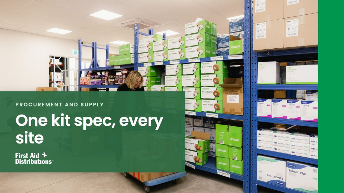 One kit spec, every site: a staff member in a warehouse aisle lined with blue racking stacked with boxed tape and wound care stock, First Aid Distributions