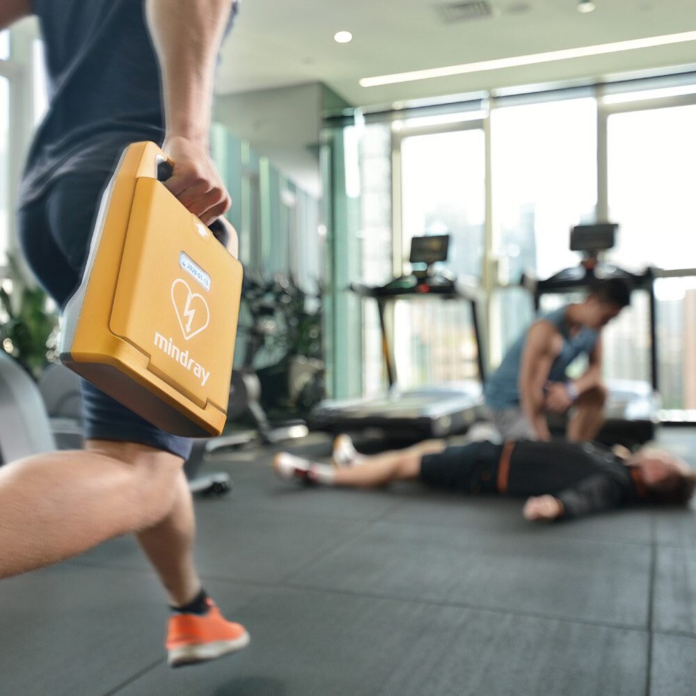 Person carrying the Mindray BeneHeart C1A AED by its handle towards a collapsed man in a gym